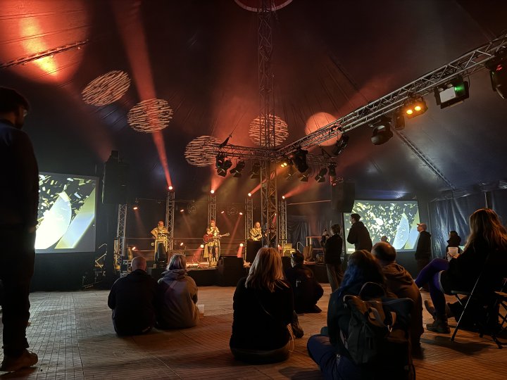 A photo of a stage in a large tent, on which three band members are performing, pressed in mokish gold robes. The left most is playing a laptop (doing the visualisations that are projected either side of the stage), in the middle is a woman singing whilst playing a Gibson SG guitar, and on the right a man is playing bass guitar. In front of the stage is a small chill crowd enjoying the show.