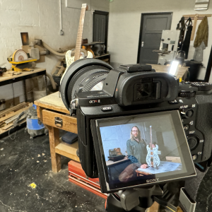 A photo of the back of a camera set up on a tripod in a workshop, pointing at a guitar on a bench. On the camera rear screen is me holding the guitar, as taken a moment ago.