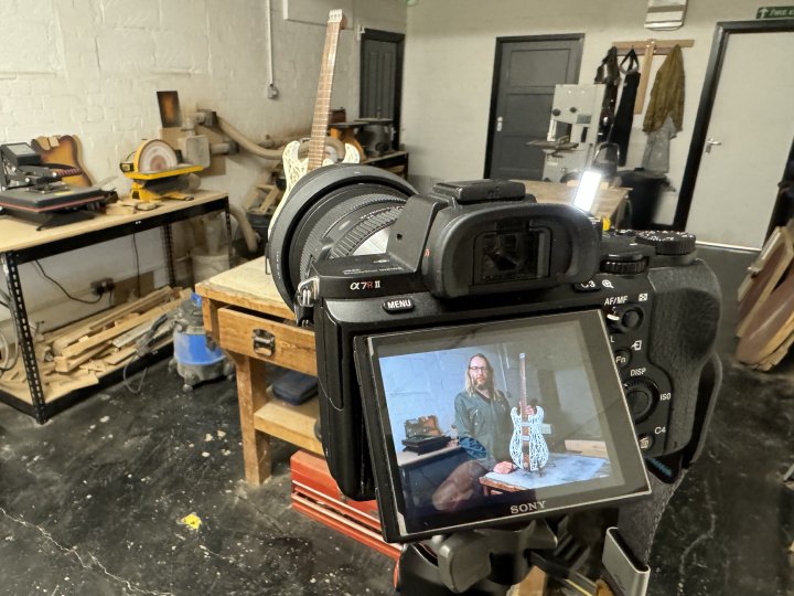 A photo of the back of a camera set up on a tripod in a workshop, pointing at a guitar on a bench. On the camera rear screen is me holding the guitar, as taken a moment ago.