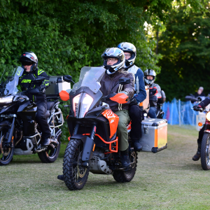 A photo of two people on a motorcycle driving along a grass lane next to a wall of trees, with a few other motorcycles near by. The main bike is a big KTM adventure bike, on which is my and Laura.