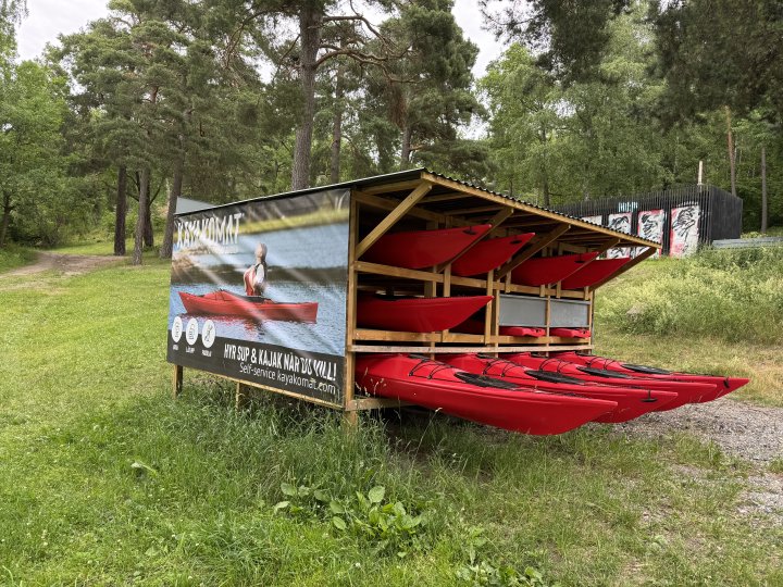 A photo of a small wooden structure in a wooded park area that houses a bunch of kayaks and stand up paddel boards. On the side is a sign that says 'kayakomat' with instructions on now to rent the kayaks.