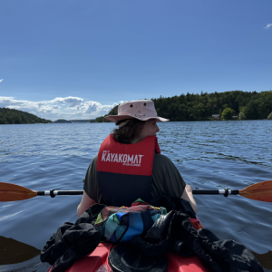 A photo taken from the back seat of a two person kayak, looking at Laura piloting up front. Above us a blue sky with a few clouds, below is blue water of lake Mälaren, and either side are forests of pine trees.