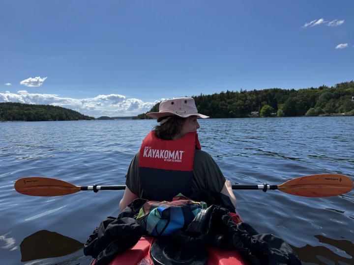 A photo taken from the back seat of a two person kayak, looking at Laura piloting up front. Above us a blue sky with a few clouds, below is blue water of lake Mälaren, and either side are forests of pine trees.