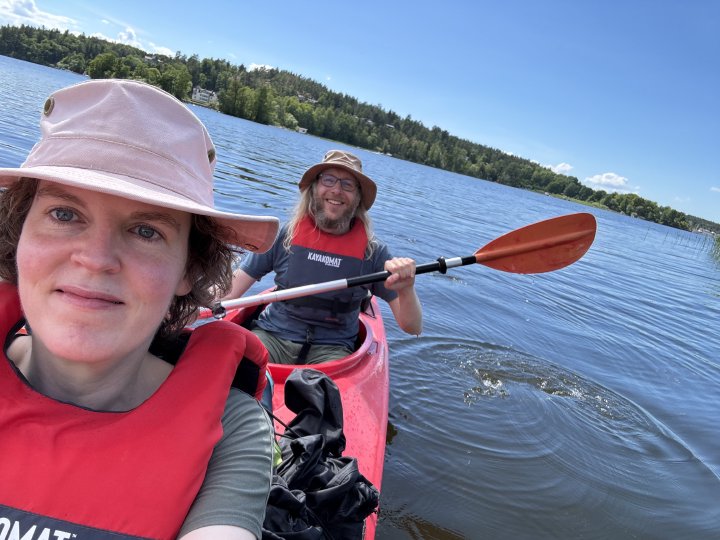 A photo taken by Laura from the front of the Kayak looking back so you can see her smiling and me in the back holding my oar, and behind me is the water of the lake, the forest of one of the islands in the lake (Fågelön) and the blue sky.