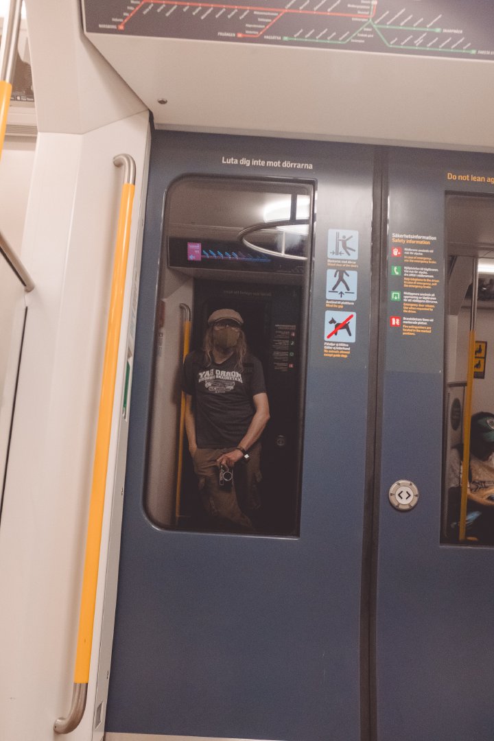 A photo of the doors of a subway train from inside the train. Reflected in the window of the door you see me holding a camera next to my leg. I'm wearing a face mask, a flat cap, and a t-shirt and cargo pants.