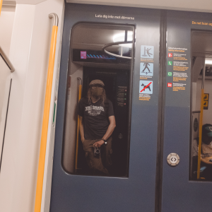A photo of the doors of a subway train from inside the train. Reflected in the window of the door you see me holding a camera next to my leg. I'm wearing a face mask, a flat cap, and a t-shirt and cargo pants.