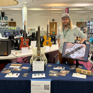 A photo of me stood in a library behind a table with several guitars, including one where the body is 3D printed.