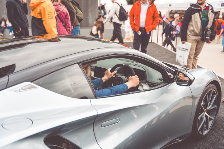 A photo of someone with a graying beard (me apparently) sat in the drivers seat of a Lotus Emira sports car park on a stand surrounded by people looking on.