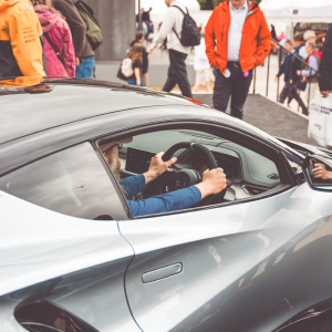 A photo of someone with a graying beard (me apparently) sat in the drivers seat of a Lotus Emira sports car park on a stand surrounded by people looking on.