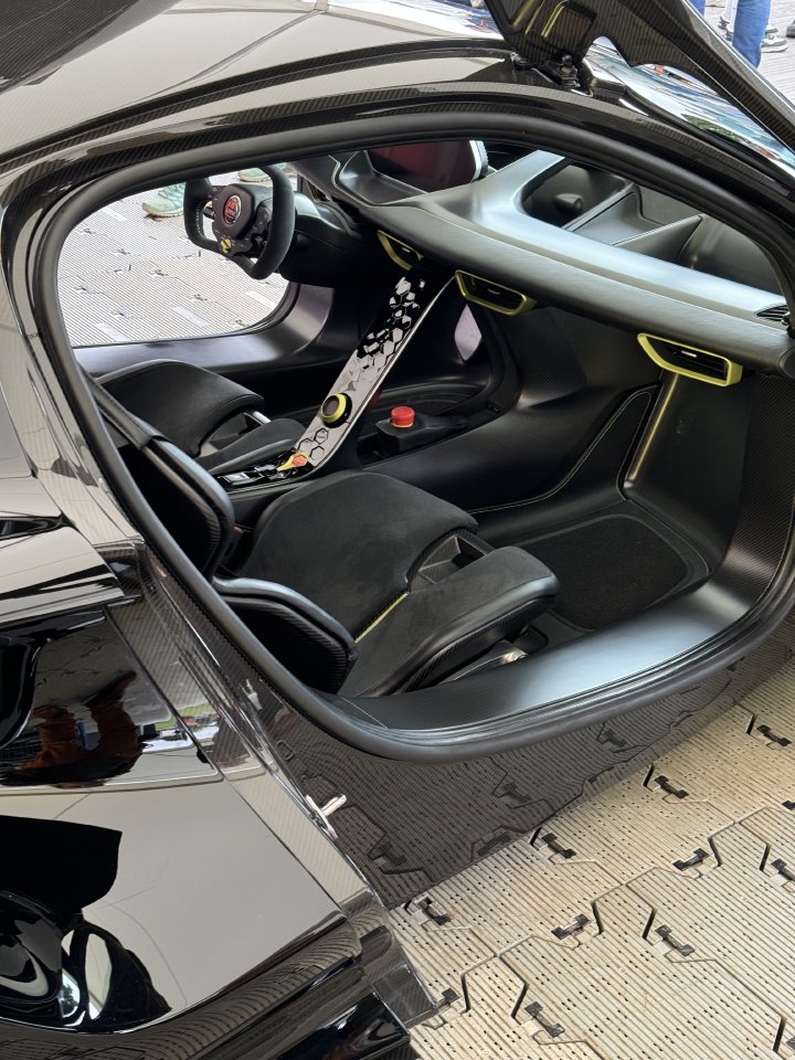 A photo of the cabin of the Lotus Evija hyper car, in which you can see it has carbon-fibre bucket seats, an F1 style racing streeing wheel, and a very minimal/stylised center console. At the bottom of the centre console, looking somewhat conspicuous against the black interior, is a standard red button on yellow emergancy stop button as seen in workshops everywhere.