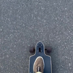 A video taken from the perspective of someone on a longboard, opening with a shot of their feet on the board going along a long pavement, then moving up to see where they are going, which is a long a pathway surrounded on both sides by tall grass, and to the left if a wheat field and the right trees.