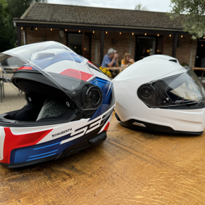 A photo of two motorcycle helmets looking away from each other on an outdoors table at a cafe. Both have the same profile, but one has white, blue and red graphics and has "S3" and "SCHUBERTH" written on it, and the other is just plain white. On the coloured one you can also make out intercom controls on the left.