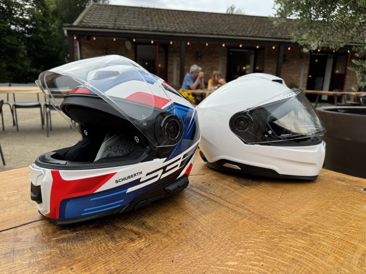 A photo of two motorcycle helmets looking away from each other on an outdoors table at a cafe. Both have the same profile, but one has white, blue and red graphics and has "S3" and "SCHUBERTH" written on it, and the other is just plain white. On the coloured one you can also make out intercom controls on the left.