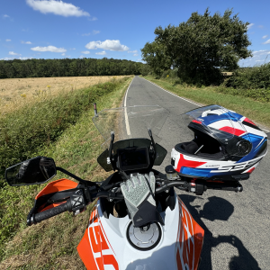 A photo taken on a bright sunny day from the view of a person sitting on a motorbike, with their gloves and helmet on the handlebars, looking down a section of single-track road with fields either side that vanishes into some woodlands.