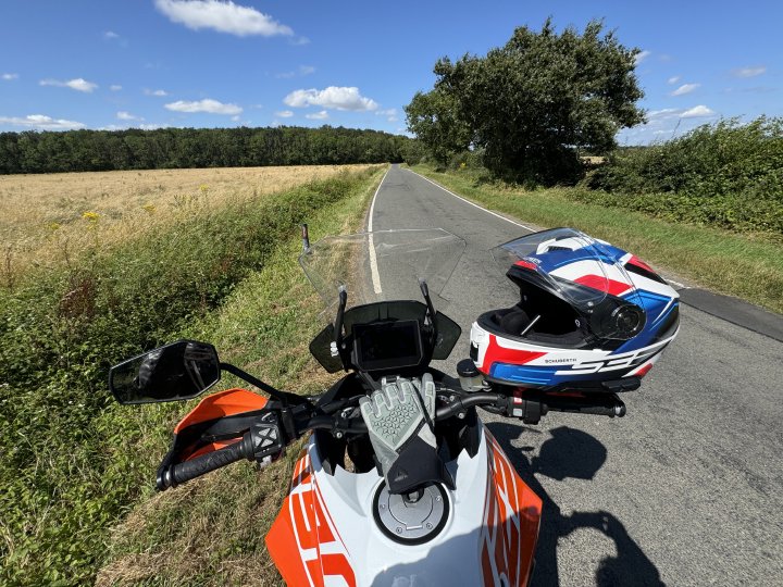 A photo taken on a bright sunny day from the view of a person sitting on a motorbike, with their gloves and helmet on the handlebars, looking down a section of single-track road with fields either side that vanishes into some woodlands.