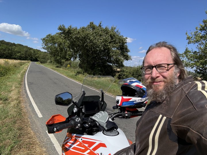 A selfie photo of me taken sitting on a motorbike holding the camera behind to show what's ahead of me, with my helmet and golves on the handlebar, and I'm squiting as the sun is in my eyes. Ahead is a section of single-track road with fields either side that vanishes into some woodlands.