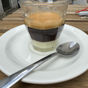 A photo of a small glass on a saucer, on a table outside a cafe. In the glass is a layered drink, where the bottom half is white, being made of condensed milk, and the top half a dark brown, being made with espresso. There is a tea spoon on the saucer also, which is for stirring the drink before you sip it.