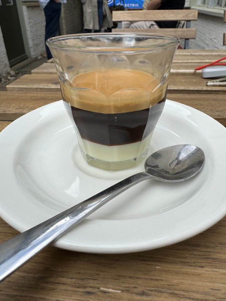 A photo of a small glass on a saucer, on a table outside a cafe. In the glass is a layered drink, where the bottom half is white, being made of condensed milk, and the top half a dark brown, being made with espresso. There is a tea spoon on the saucer also, which is for stirring the drink before you sip it.