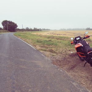 Ett foto av en stora orange KTM motorcykel nara vägen det gå genom en plat gård landskap. Himlen är grå och det är lite dimmigt