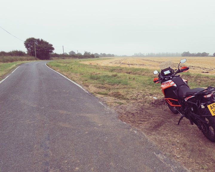 Ett foto av en stora orange KTM motorcykel nara vägen det gå genom en plat gård landskap. Himlen är grå och det är lite dimmigt