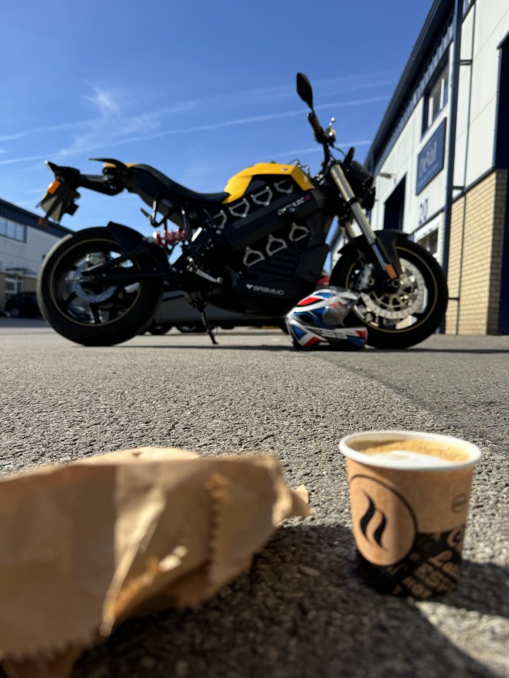 En foto från marken av an svart och gula el-motorcykel, parkerad med hjälmen på marken bredvid. Nara kamera är en litet kaffe.