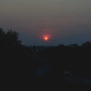 A photo of the sun about set over a city skyline, the sun is a distinct deep red circle in a hazy sky.