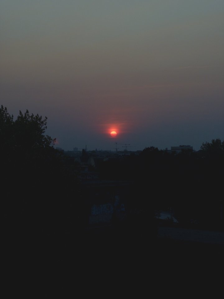 A photo of the sun about set over a city skyline, the sun is a distinct deep red circle in a hazy sky.