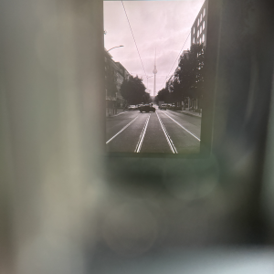 A photo of a photo of a look down a Berlin street with the tall spire of the TV tower at the end of it. The inner photo is offset to the top right somewhat, the frame around it is all out of focus lines (it is in fact the eye finder of a camera that is being photographed)