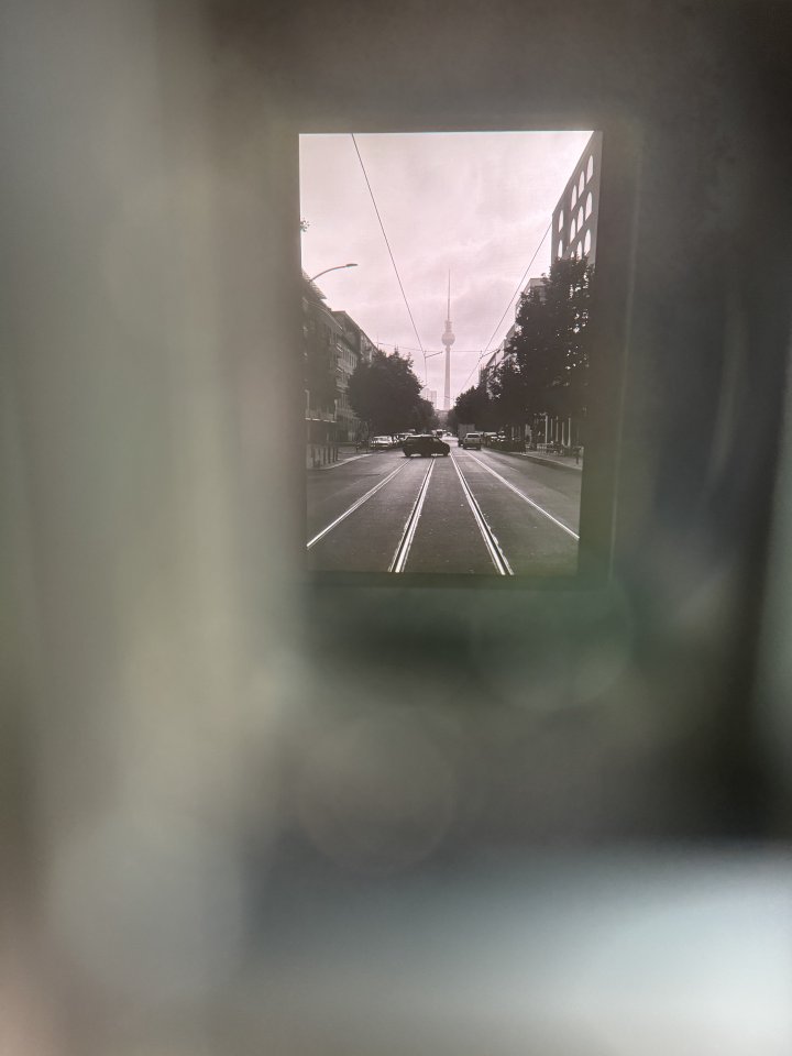 A photo of a photo of a look down a Berlin street with the tall spire of the TV tower at the end of it. The inner photo is offset to the top right somewhat, the frame around it is all out of focus lines (it is in fact the eye finder of a camera that is being photographed)