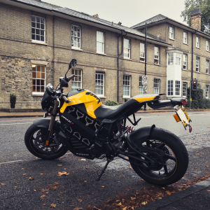 A photo of a bright yellow and black electric motorbike parked on a damp looking road with a weather brick building (Darwin College) in the background, and autumn fallen leaves on the ground in the foreground.