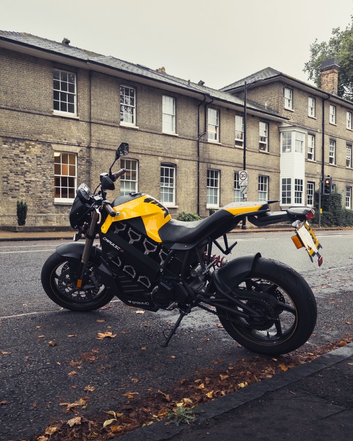 A photo of a bright yellow and black electric motorbike parked on a damp looking road with a weather brick building (Darwin College) in the background, and autumn fallen leaves on the ground in the foreground.