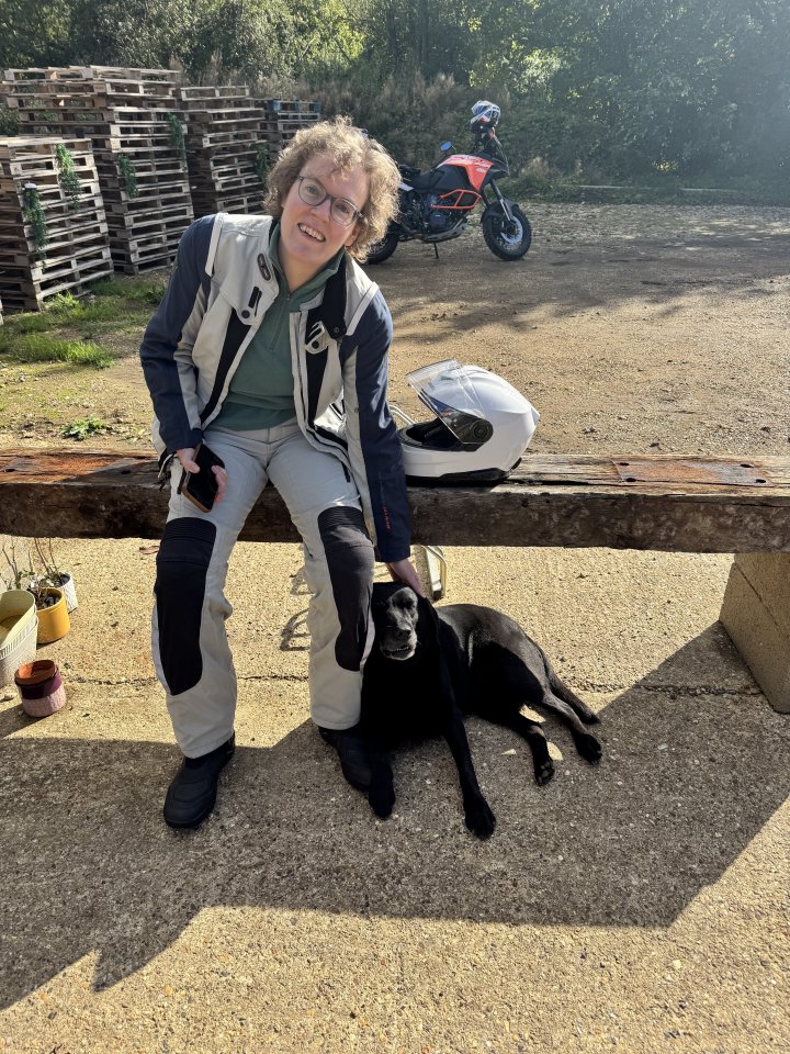 A photo of Laura sat on a bench in her motorcycle outfit, and lent against her leg is a black labrador dog, as if it's known her its entire life (they met about two minutes earlier). In the background sits the bright orange of a large KTM adventure motorbike.