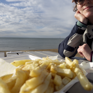 A photo of a sunlit Laura in motorcycle gear and a cap, sat at a table on which sit some chips, and behind her is the sea.