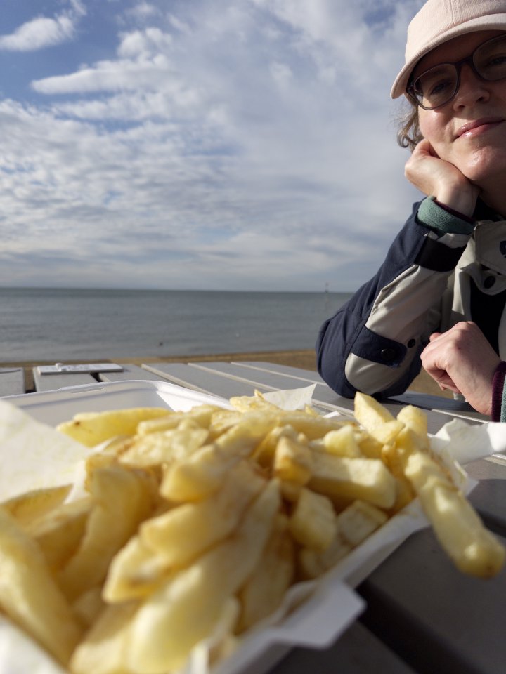 A photo of a sunlit Laura in motorcycle gear and a cap, sat at a table on which sit some chips, and behind her is the sea.