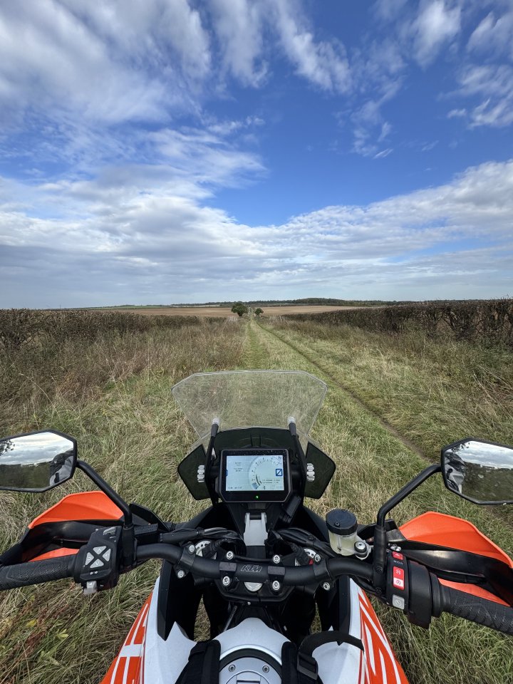 A photo taken from the seat of a KTM orange and white large adventure motorcycle, looking down a green lane set between two fields, running off straight ahead into the distance. Above the sky is bright blue with clouds in it.