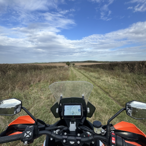 A photo taken from the seat of a KTM orange and white large adventure motorcycle, looking down a green lane set between two fields, running off straight ahead into the distance. Above the sky is bright blue with clouds in it.