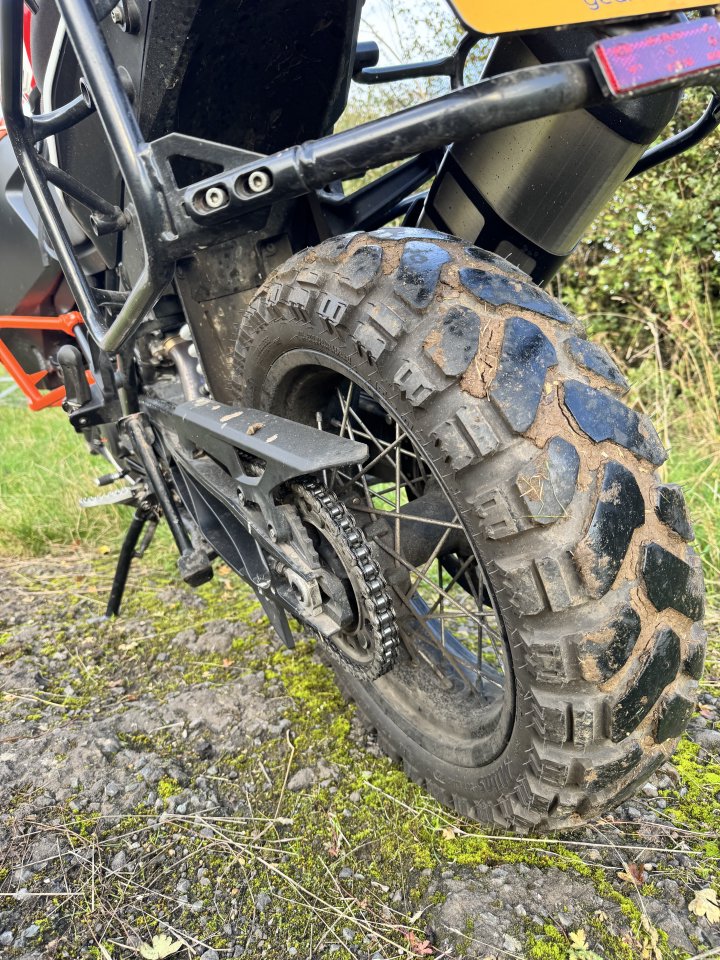 A close up photo of the rear tyre of a motorbike, showing the chunky tread pattern with an inch between blocks, and much of that pattern filled with mud.