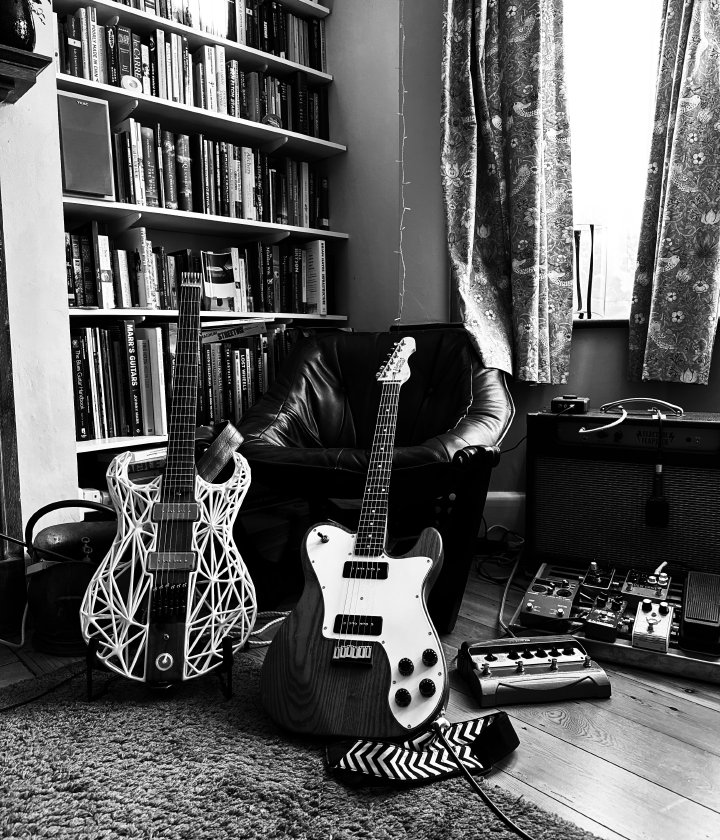 A black and white photo of a pair of electric guitars sat in a living room, one in a stand another lent against a chair. One side is a set of book shelves, and on the other side an amp and a small pedal board.