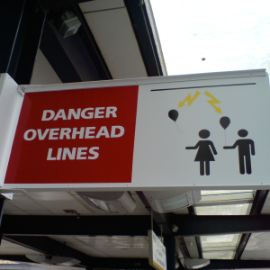 An overhead sign from a train platform showing two children with balloons and lightning striking their balloons from a line above, with the words 'DANGER OVERHEAD LINES'