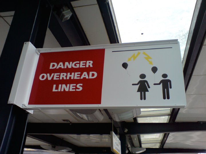 An overhead sign from a train platform showing two children with balloons and lightning striking their balloons from a line above, with the words 'DANGER OVERHEAD LINES'