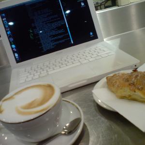 A photo of my laptop in a cafe with a cappuccino and croissant next to it.