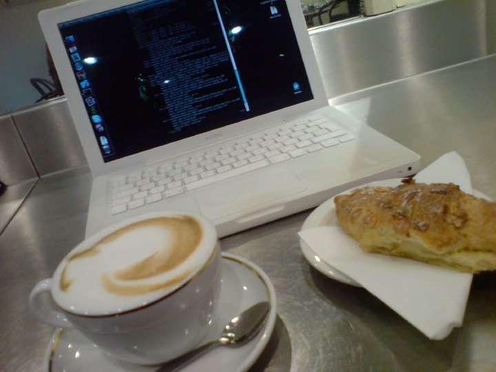 A photo of my laptop in a cafe with a cappuccino and croissant next to it.