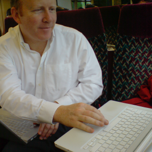Quantin sat on a train with a MacBook Pro on his knees whilst typing on a MacBook on the table.