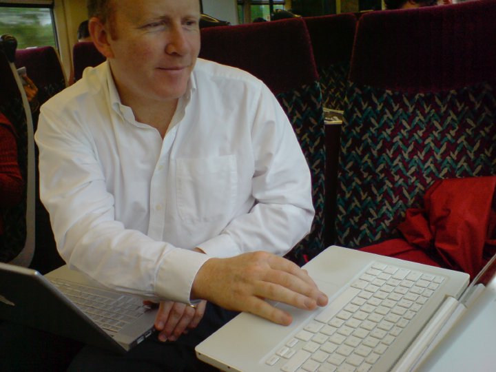 Quantin sat on a train with a MacBook Pro on his knees whilst typing on a MacBook on the table.