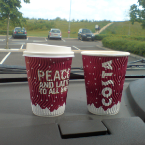 Two costa coffee cups sit on the dashboard of a car, one says 'COSTA' on it, and the other 'PEACE AND LATTES TO ALL MEN'