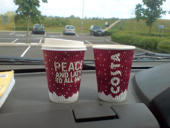 Two costa coffee cups sit on the dashboard of a car, one says 'COSTA' on it, and the other 'PEACE AND LATTES TO ALL MEN'