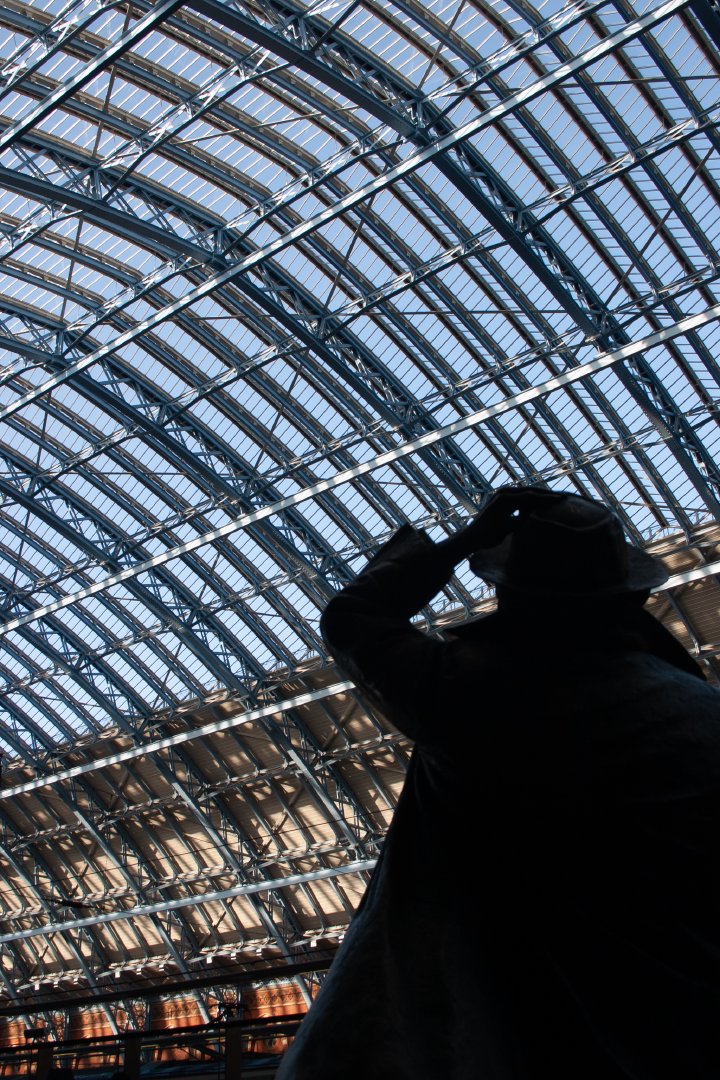 A shot looking at the glass and iron roof of St Pancras station over the sholder of a man clutching his hat.