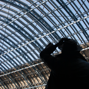 A shot looking at the glass and iron roof of St Pancras station over the sholder of a man clutching his hat.