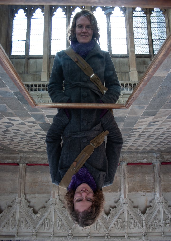 A photo of Laura looking into a mirror laid on a table in Ely Cathedral, to make it easier to see the ceiling. The photo is looking at Laura in the mirror, and the whole picture has been flipped to mirror Laura is the right way up, and non-mirror Laura is where the relfection should be.